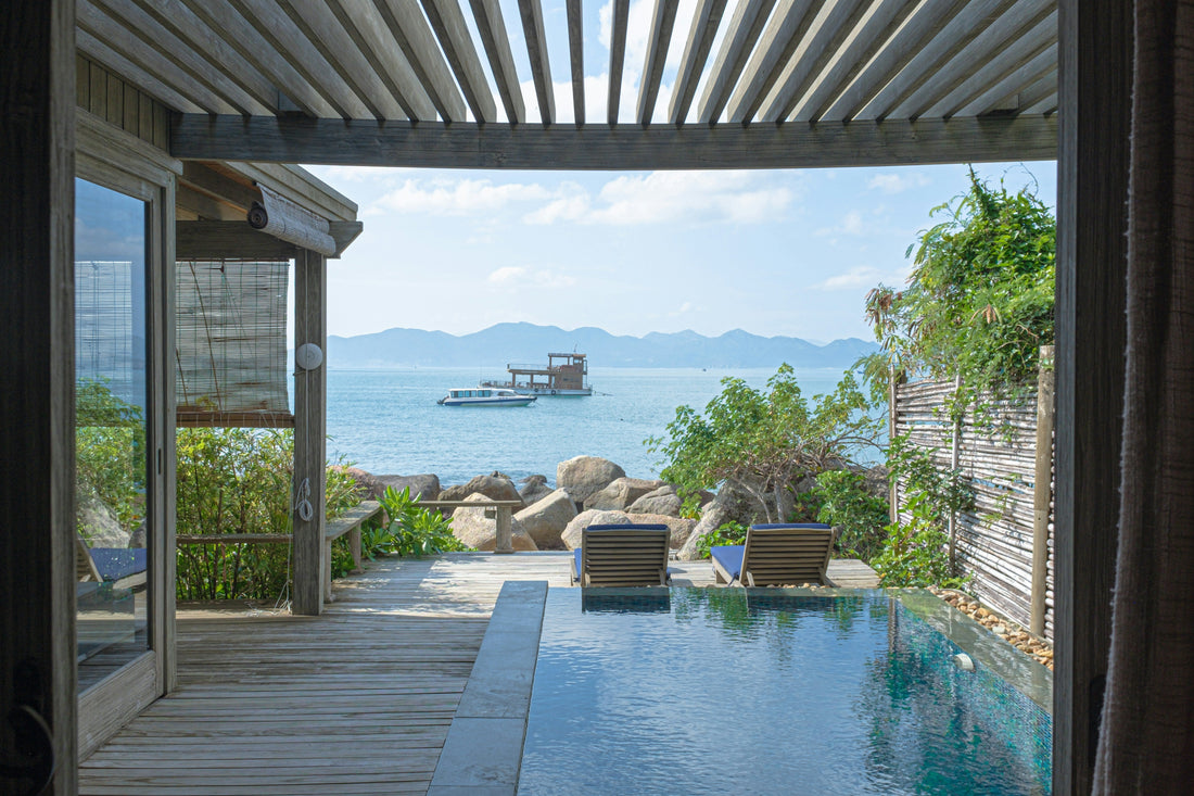 Two wooden lounge chairs with blue cushions face the water, separated from the sea by large rocks. 