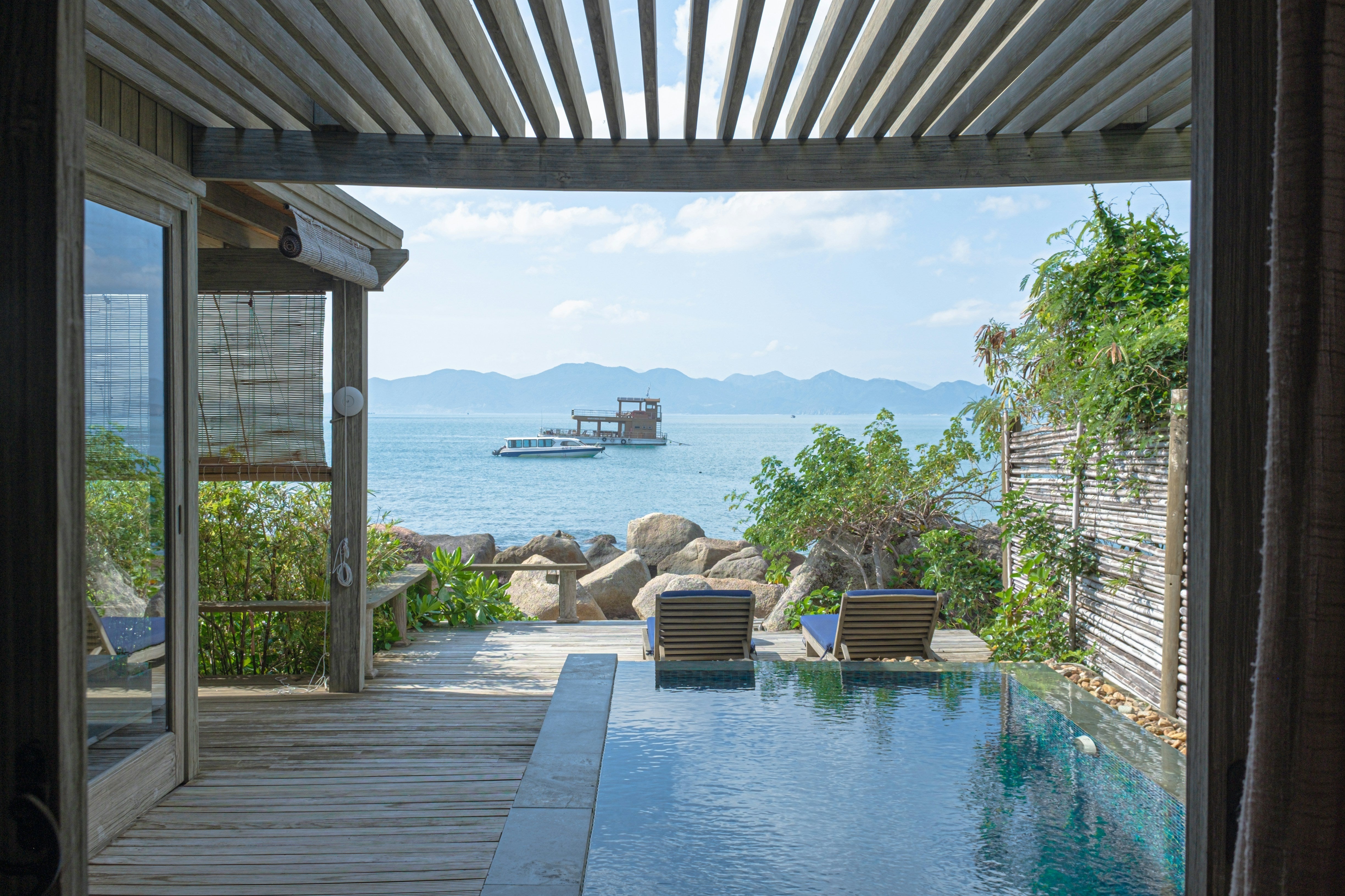 Two wooden lounge chairs with blue cushions face the water, separated from the sea by large rocks. 